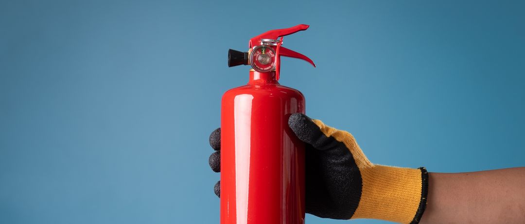 Eine Person mit gelben und schwarzen Handschuhen hält einen roten Feuerlöscher mit rotem Griff. Der Hintergrund ist blau.
