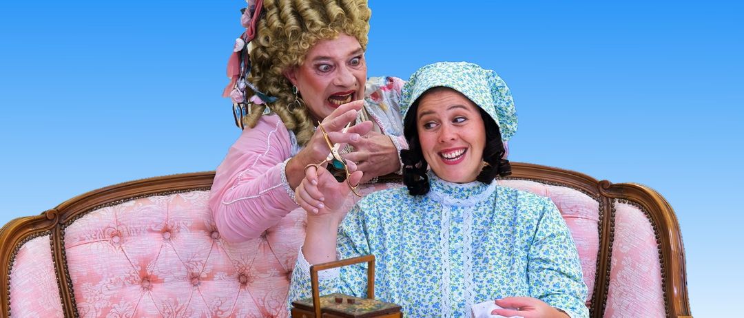 Zwei Frauen sitzen auf einem rosa Sofa, eine hält eine Schere, die andere ein Tuch. Ein Holzkasten steht auf einem Tisch vor ihnen.