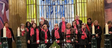 Ein Chor erwachsener Menschen mit roten Schals steht vor einem geschmückten Altar, mit Kerzen und Blumen auf der Bühne, einem farbenfrohen Buntglasfenster dahinter.
