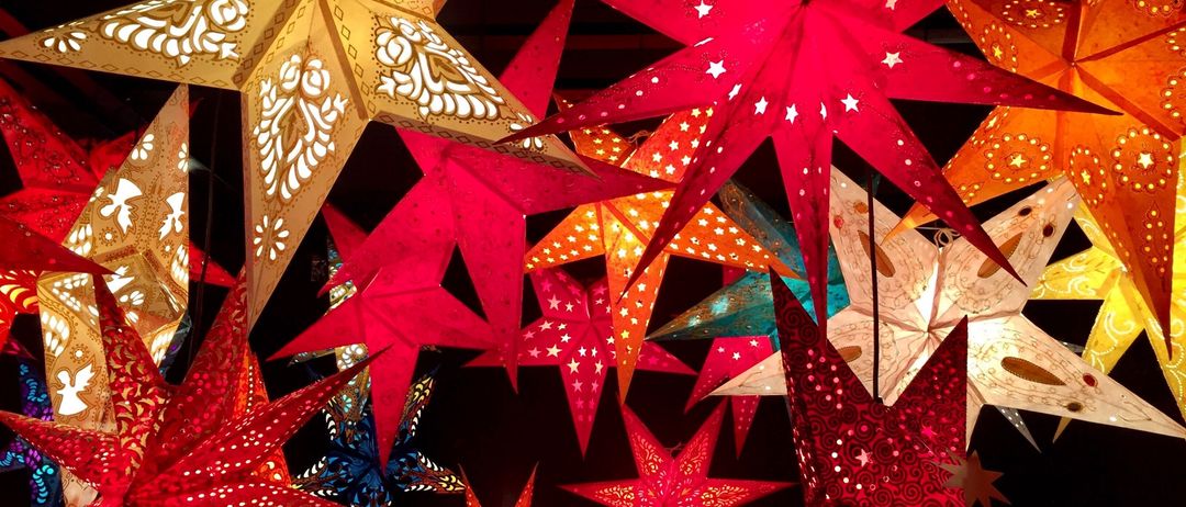 Verschiedene Papiersterne in leuchtenden Farben hängen von der Decke. Sie haben komplizierte Muster und Lichtreflexionen, die einen festlichen Touch hinzufügen.