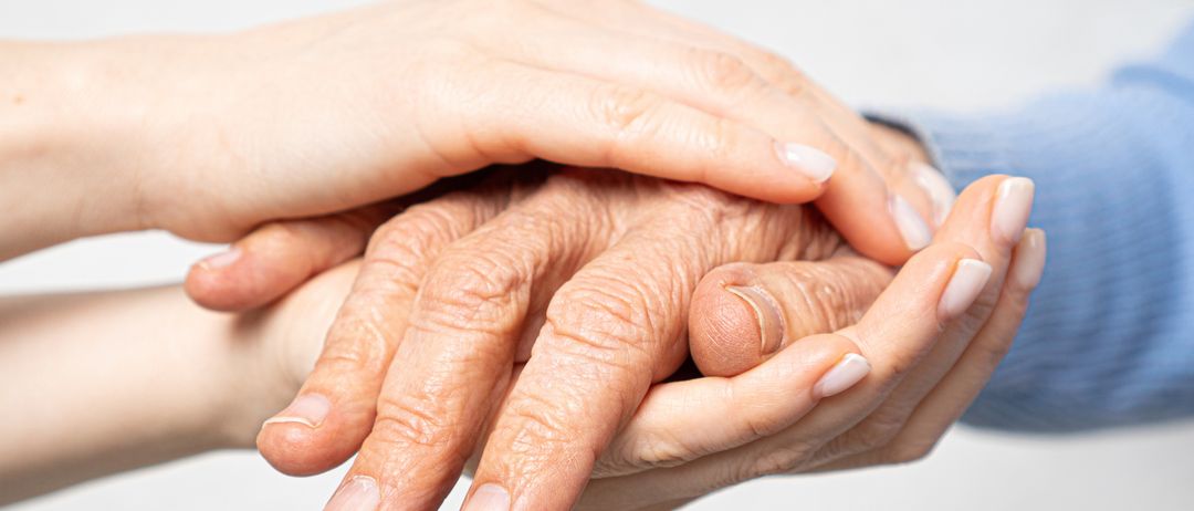 Zwei Hände halten die Hände einer älteren Person und zeigen Fürsorge und Unterstützung. Die Hände sind faltig und gealtert, mit weißen Nägeln.