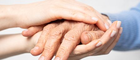 Zwei Hände halten die Hände einer älteren Person und zeigen Fürsorge und Unterstützung. Die Hände sind faltig und gealtert, mit weißen Nägeln.