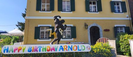 A yellow house with green shutters and a birthday sign in front of it. The sign reads 'Happy Birthday Arnold' with a statue of a muscular man in front of it.