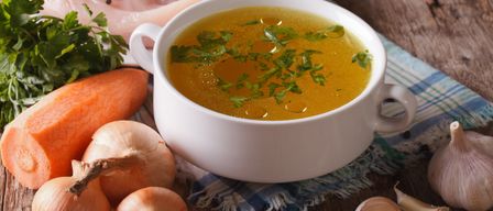 Eine Schüssel Hühnerbrühe mit Petersiliengarnitur, begleitet von einer Scheibe rohem Hühnchen, Karotten, Zwiebeln und Knoblauch auf einem Holztisch.