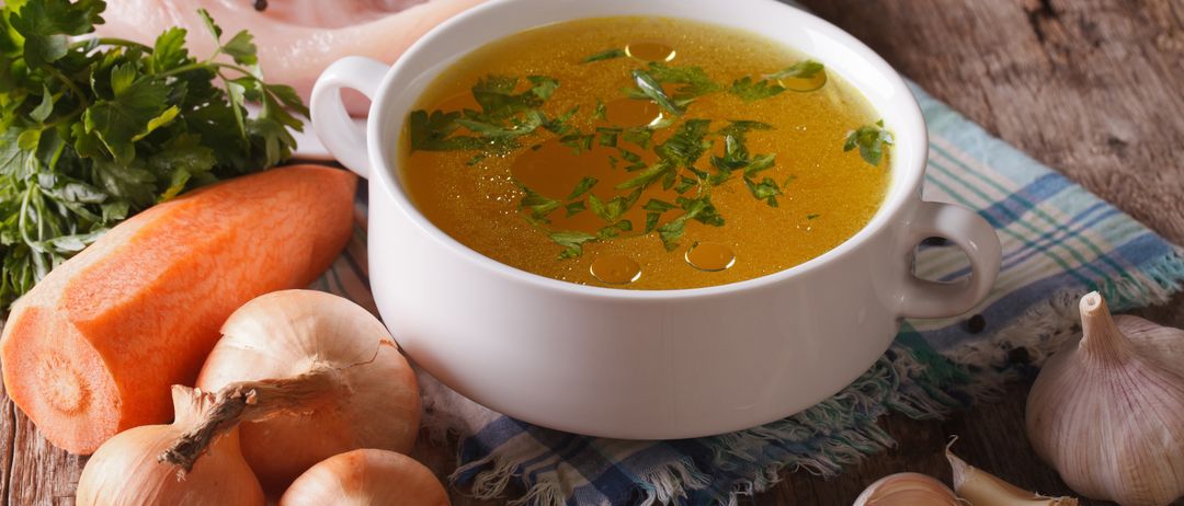 Eine Schüssel Hühnerbrühe mit Petersiliengarnitur, begleitet von einer Scheibe rohem Hühnchen, Karotten, Zwiebeln und Knoblauch auf einem Holztisch.