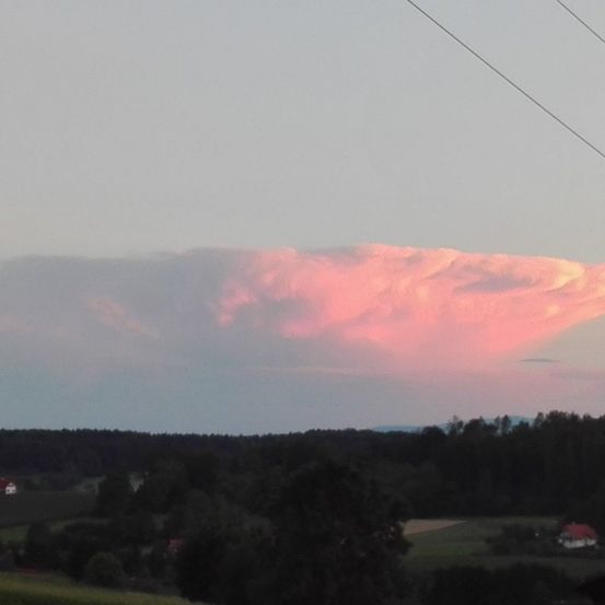 Eine ländliche Landschaft mit Bäumen, Häusern und einer großen Wolkenformation am Horizont bei Sonnenuntergang. Der Himmel ist pink, und es gibt Stromleitungen darüber.