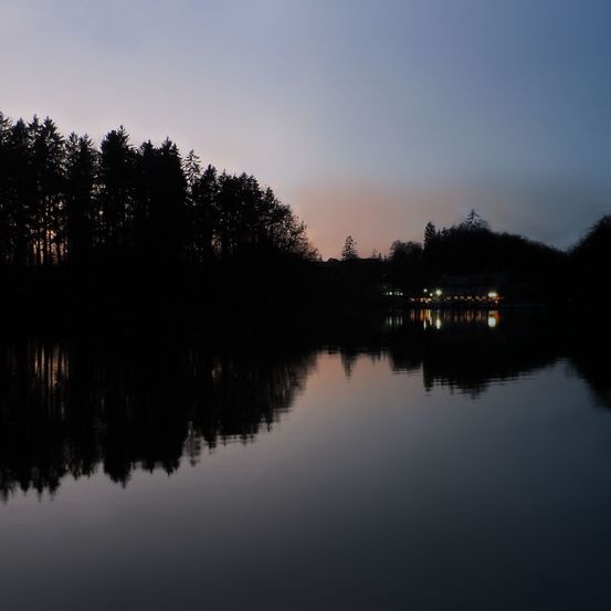 Ein ruhiger See mit silhouettierten Bäumen und entfernten Lichtern in der Dämmerung. Der Himmel ist ein sanfter Farbverlauf von Blau und Rosa.