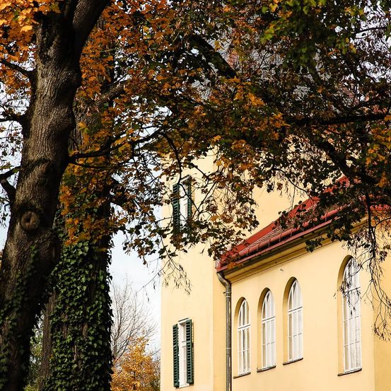 Ein Gebäude mit gelben Wänden und einem roten Dach. Das Gebäude hat Rundbogenfenster. Ein Baum mit grünen und gelben Blättern steht neben dem Gebäude.