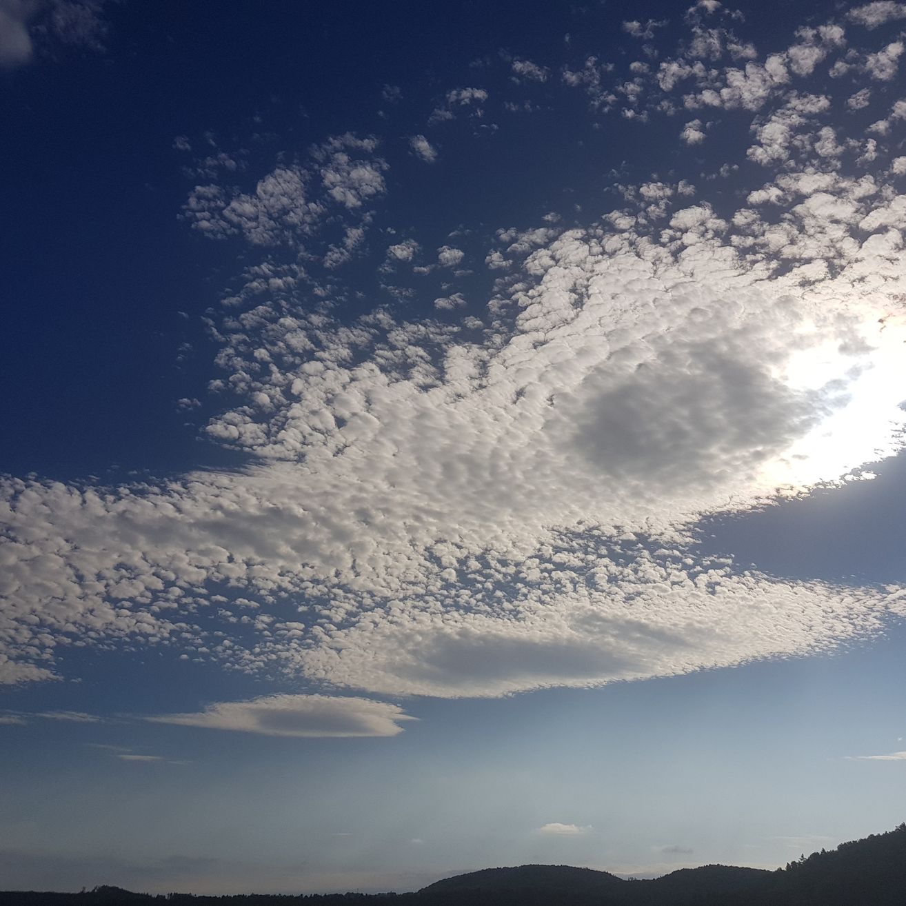 Der Himmel ist mit flauschigen Wolken gefüllt, vor einem klaren blauen Hintergrund, mit hellem Sonnenlicht. Hügel und Berge sind in der Ferne sichtbar.