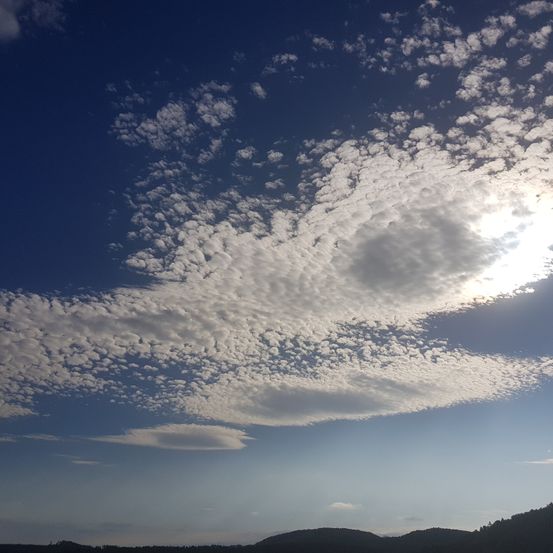 Der Himmel ist mit flauschigen Wolken gefüllt, vor einem klaren blauen Hintergrund, mit hellem Sonnenlicht. Hügel und Berge sind in der Ferne sichtbar.