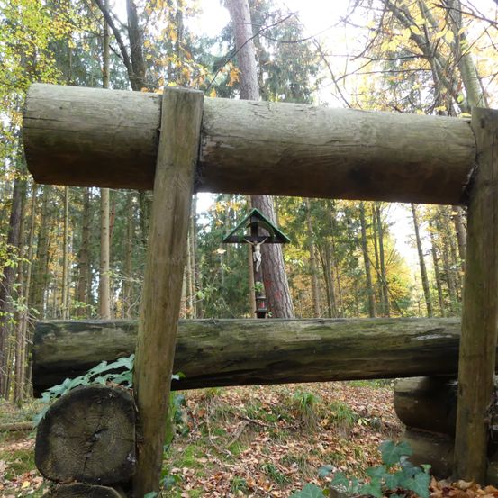 Eine rustikale Holzstruktur steht in einem Wald, mit einem Kreuz auf der Spitze und umgeben von umgestürzten Baumstämmen und gefallenen Blättern.
