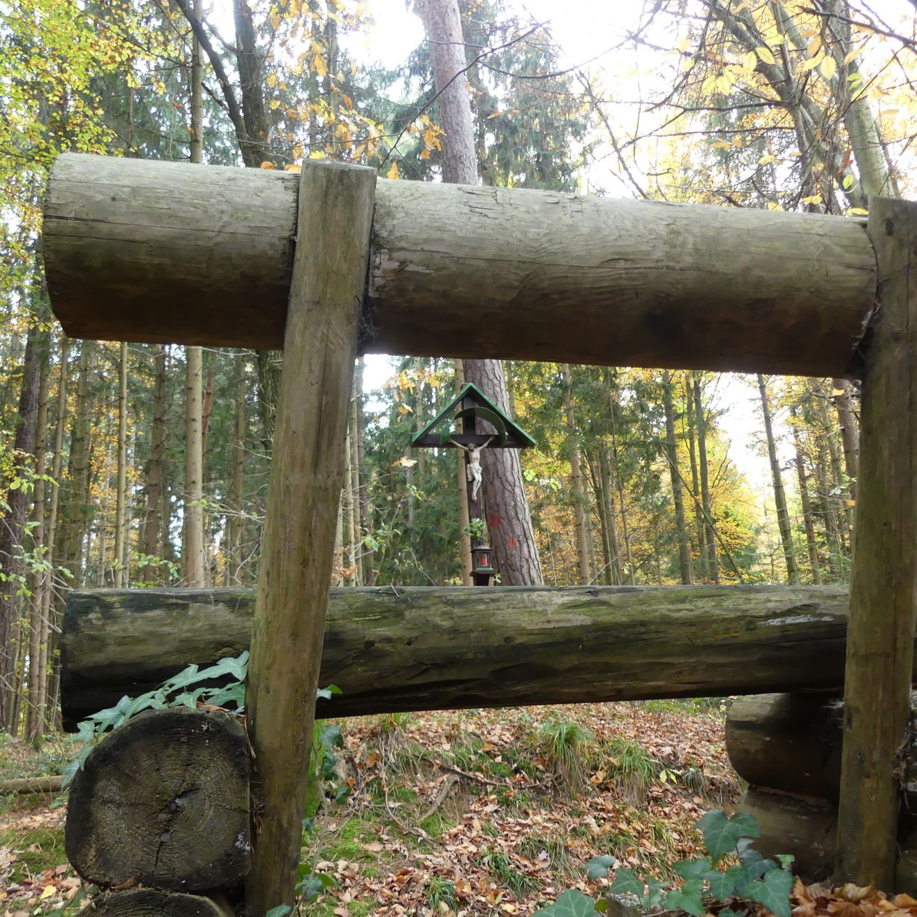 Eine rustikale Holzstruktur steht in einem Wald, mit einem Kreuz auf der Spitze und umgeben von umgestürzten Baumstämmen und gefallenen Blättern.