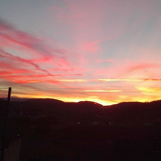 Ein Sonnenuntergang mit lebhaften rosa und gelben Farbtönen füllt den Himmel und taucht eine Berglandschaft in warmes Licht. Unten sind Umrisse von Gebäuden und Bäumen zu sehen.