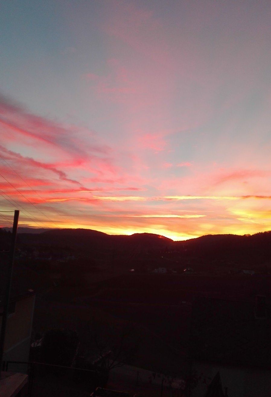 Ein Sonnenuntergang mit lebhaften rosa und gelben Farbtönen füllt den Himmel und taucht eine Berglandschaft in warmes Licht. Unten sind Umrisse von Gebäuden und Bäumen zu sehen.