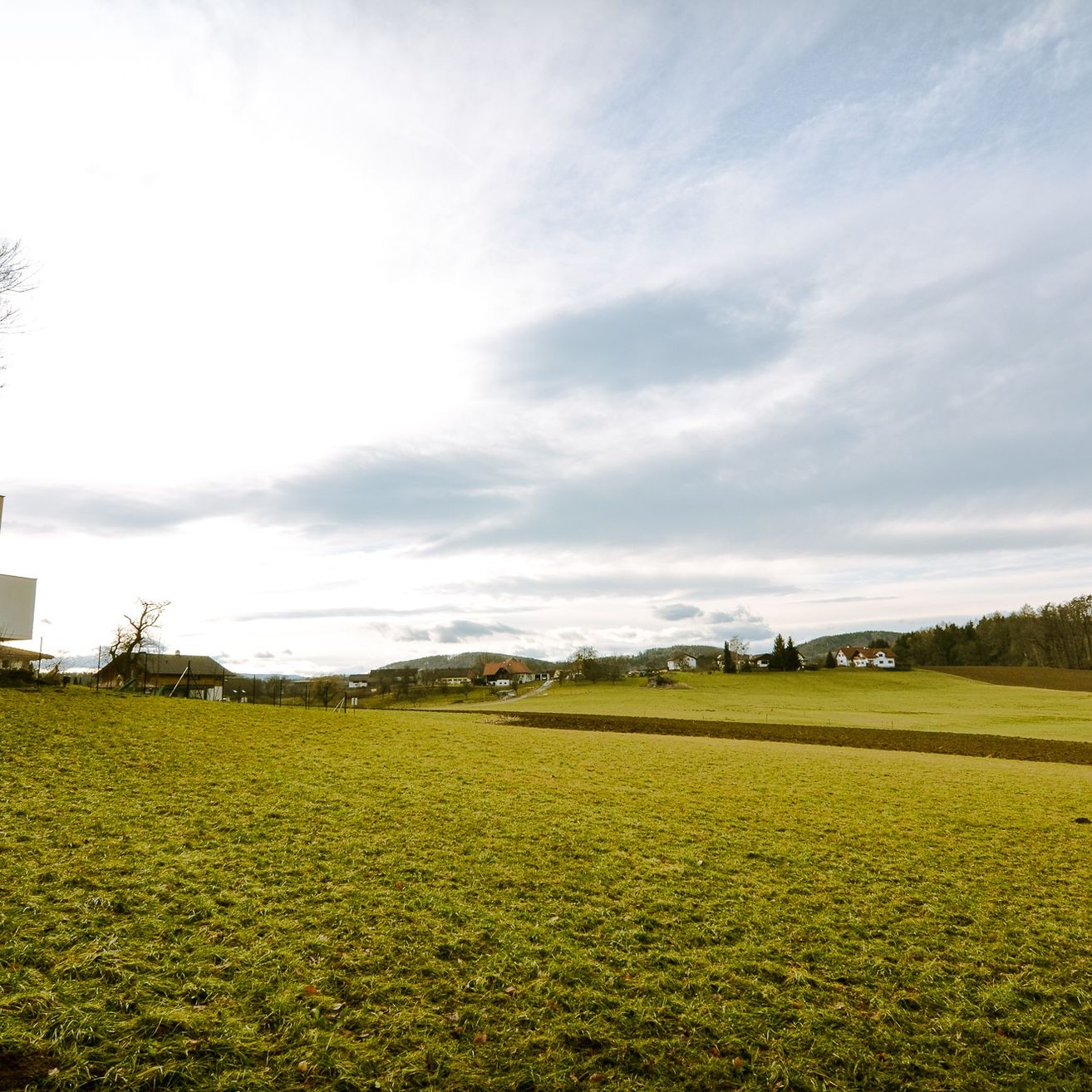 Ein grünes Feld unter einem bewölkten Himmel mit Häusern in der Ferne und einem kleinen Gebäude auf der linken Seite.