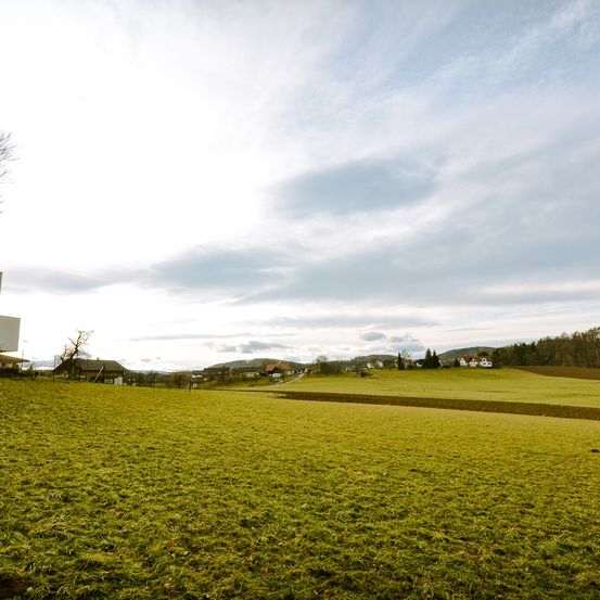 Ein grünes Feld unter einem bewölkten Himmel mit Häusern in der Ferne und einem kleinen Gebäude auf der linken Seite.