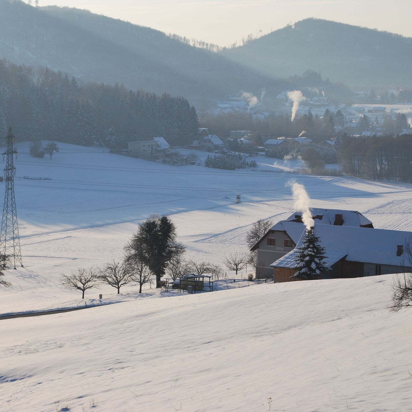 Eine verschneite Landschaft mit einem Haus und einem Schornstein, der Rauch ausstößt. In der Ferne gibt es Berge und ein Dorf. Der Himmel ist mit Nebel bedeckt.