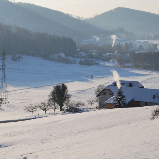 Eine verschneite Landschaft mit einem Haus und einem Schornstein, der Rauch ausstößt. In der Ferne gibt es Berge und ein Dorf. Der Himmel ist mit Nebel bedeckt.