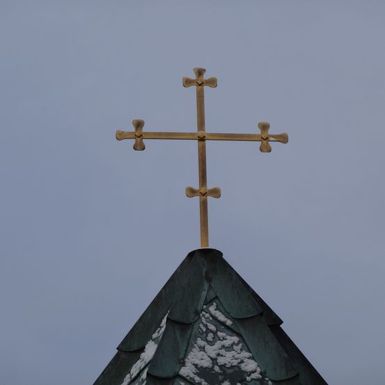 Ein goldenes Kreuz auf einem grünen Dach mit Schnee, vor einem blauen Himmel.
