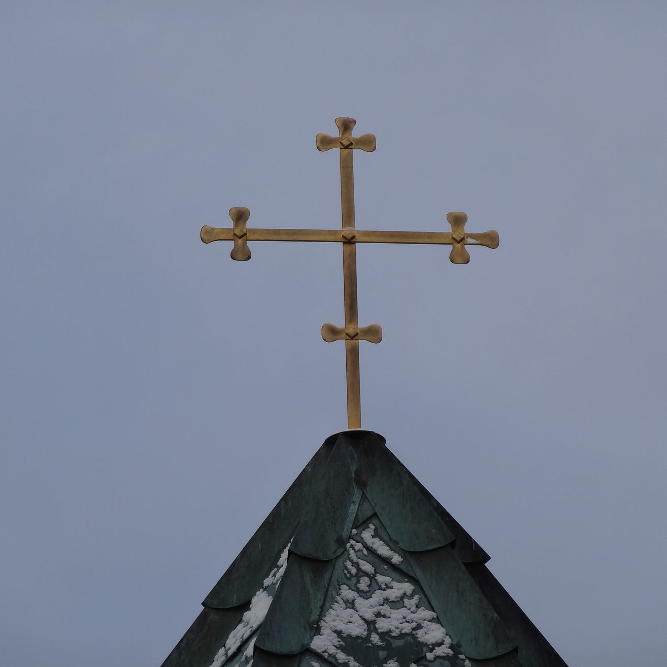 Ein goldenes Kreuz auf einem grünen Dach mit Schnee, vor einem blauen Himmel.