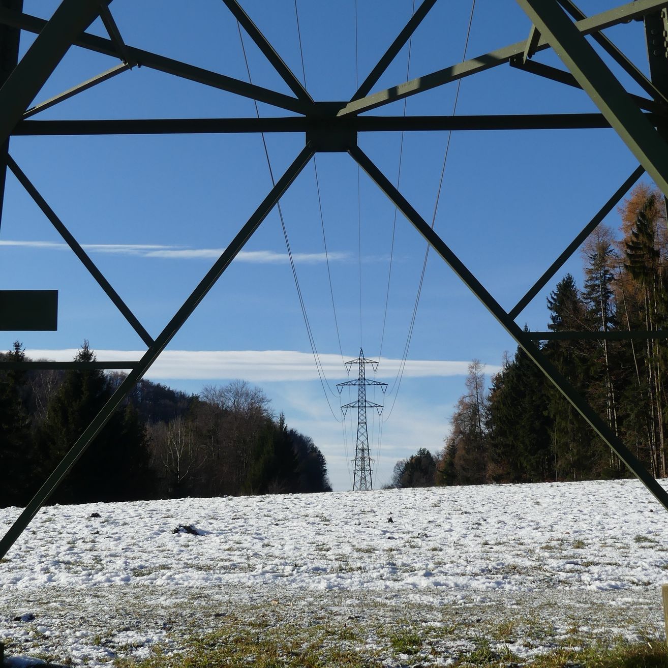 Ein Strommast steht in einem verschneiten Feld unter einem klaren blauen Himmel, eingerahmt von Metallstrukturen und umgeben von Bäumen.