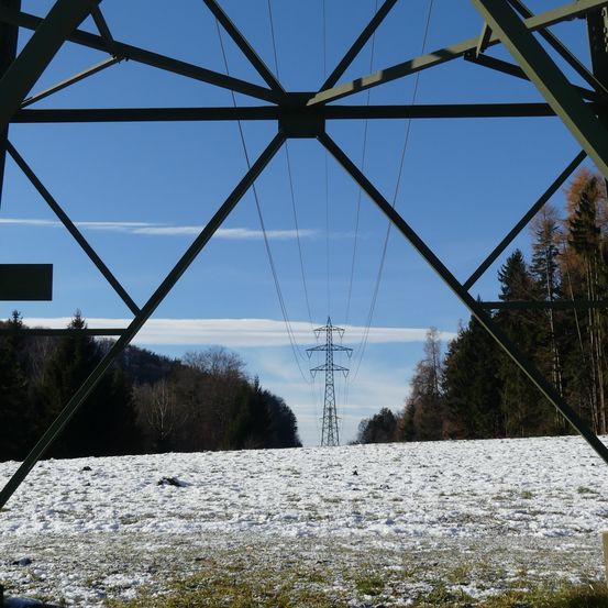 Ein Strommast steht in einem verschneiten Feld unter einem klaren blauen Himmel, eingerahmt von Metallstrukturen und umgeben von Bäumen.