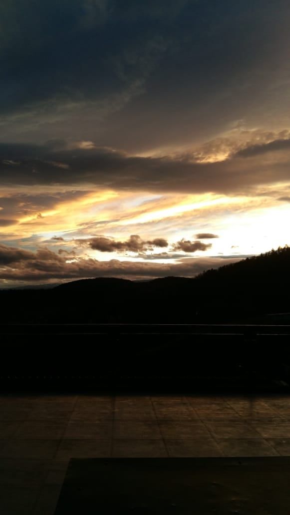 Sonnenuntergang mit goldenen Strahlen über einem Gebirgszug und dunklen Wolken, vom Balkon aus gesehen.