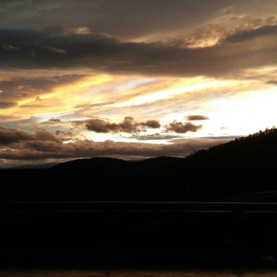 Sonnenuntergang mit goldenen Strahlen über einem Gebirgszug und dunklen Wolken, vom Balkon aus gesehen.