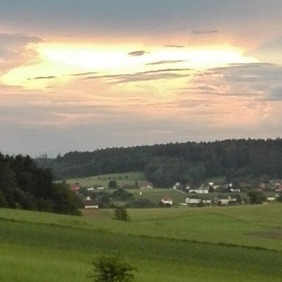 Eine ländliche Landschaft mit einer leuchtenden Sonne am Himmel, die ein grünes Feld und entfernte Häuser beleuchtet.