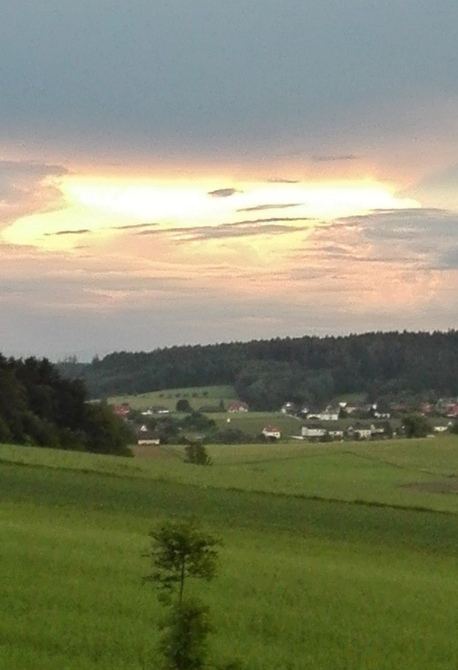 Eine ländliche Landschaft mit einer leuchtenden Sonne am Himmel, die ein grünes Feld und entfernte Häuser beleuchtet.