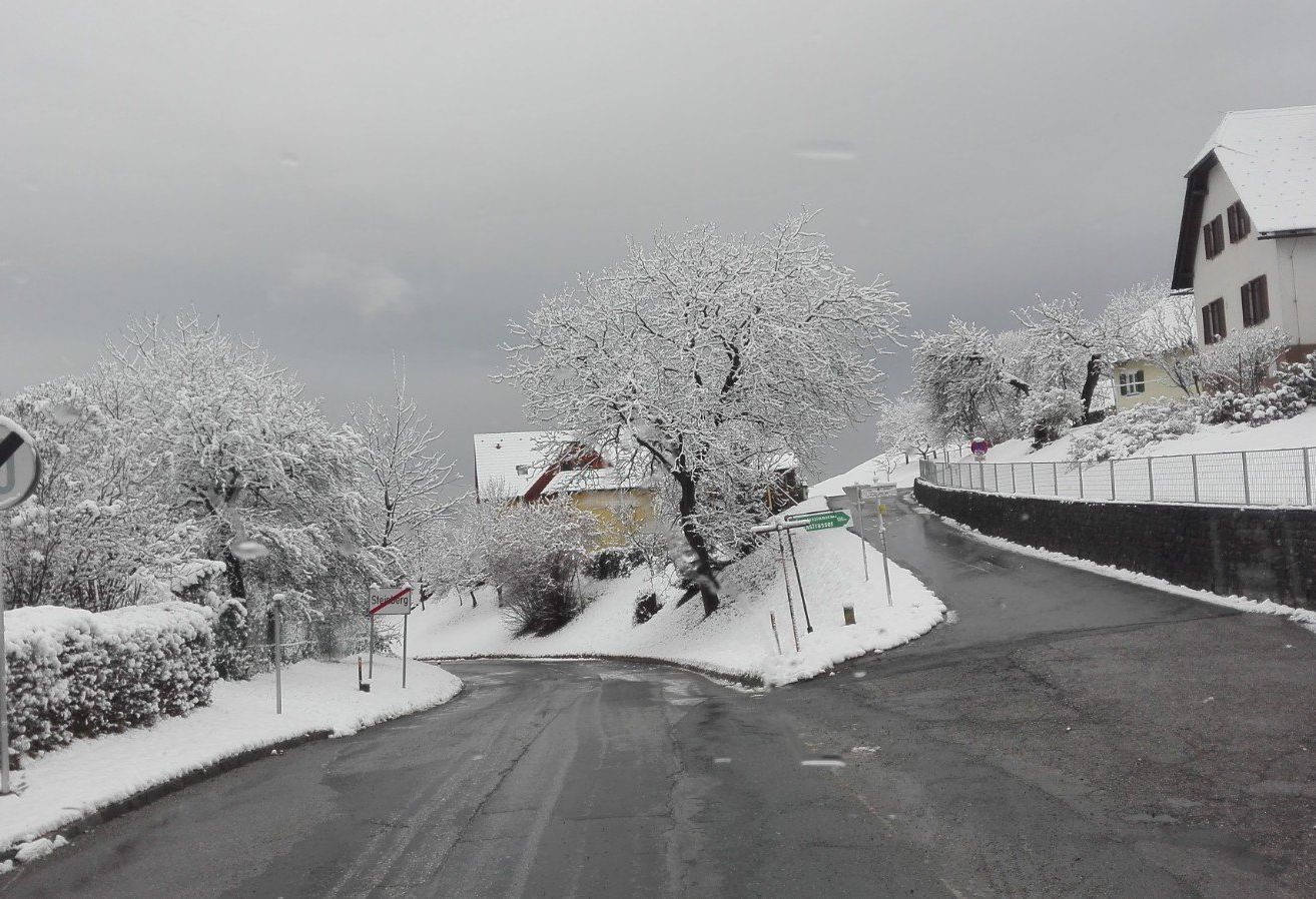 Eine verschneite Straße biegt nach links, mit einem gelben Haus in der Ferne. Schneebedeckte Bäume säumen die Seiten, und ein Straßenschild weist nach links. Der Himmel ist bedeckt.