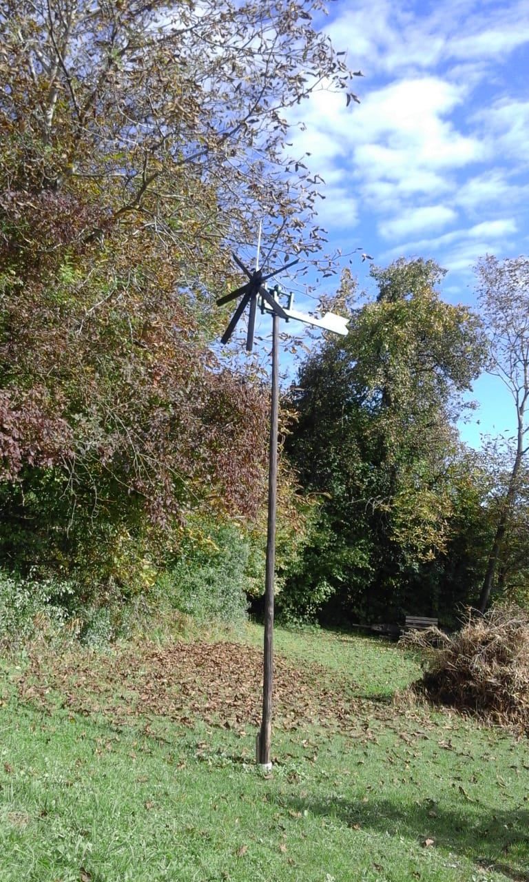 Eine Windmühle steht in einem Feld, umgeben von Herbstblättern und Bäumen. Die Windmühlenflügel drehen sich, und ein Holzpfahl stützt sie. Der Himmel ist klar mit einigen Wolken.