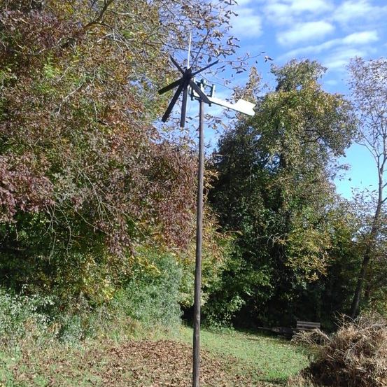 Eine Windmühle steht in einem Feld, umgeben von Herbstblättern und Bäumen. Die Windmühlenflügel drehen sich, und ein Holzpfahl stützt sie. Der Himmel ist klar mit einigen Wolken.