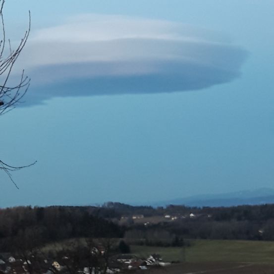 Eine große, weiße Wolke schwebt über einer Landschaft mit grünen Feldern und Bäumen. Der Himmel ist blau.