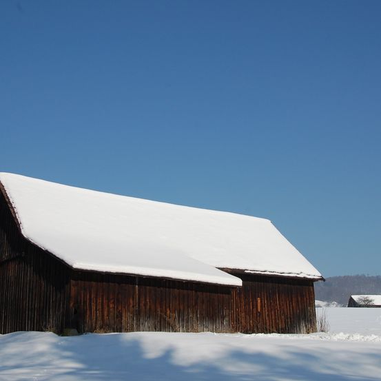 Ein hölzerner Scheunenbau mit schneebedecktem Dach steht allein in einem verschneiten Feld unter einem klaren blauen Himmel.