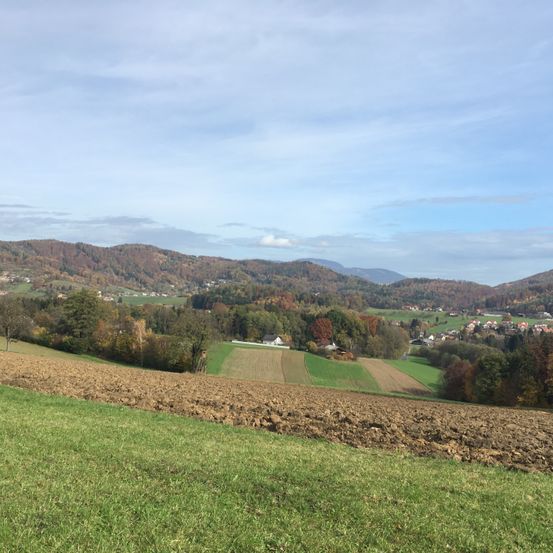 Ein Panoramablick auf eine ländliche Landschaft mit sanften Hügeln und einem gepflügten Feld unter einem teilweise bewölkten Himmel.