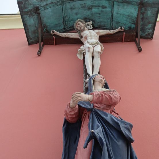 Eine Statue einer Frau mit einem blauen Umhang kniet unter einem Kruzifix an einer roten Wand. Das Kreuz ist grün mit Metallstützen.