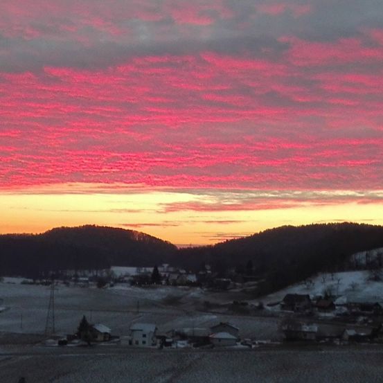 Ein Sonnenuntergangsblick auf eine verschneite Landschaft mit Bergen in der Ferne und einem kleinen Dorf darunter. Der Himmel ist in Rot-, Orange- und Gelbtönen gemalt.