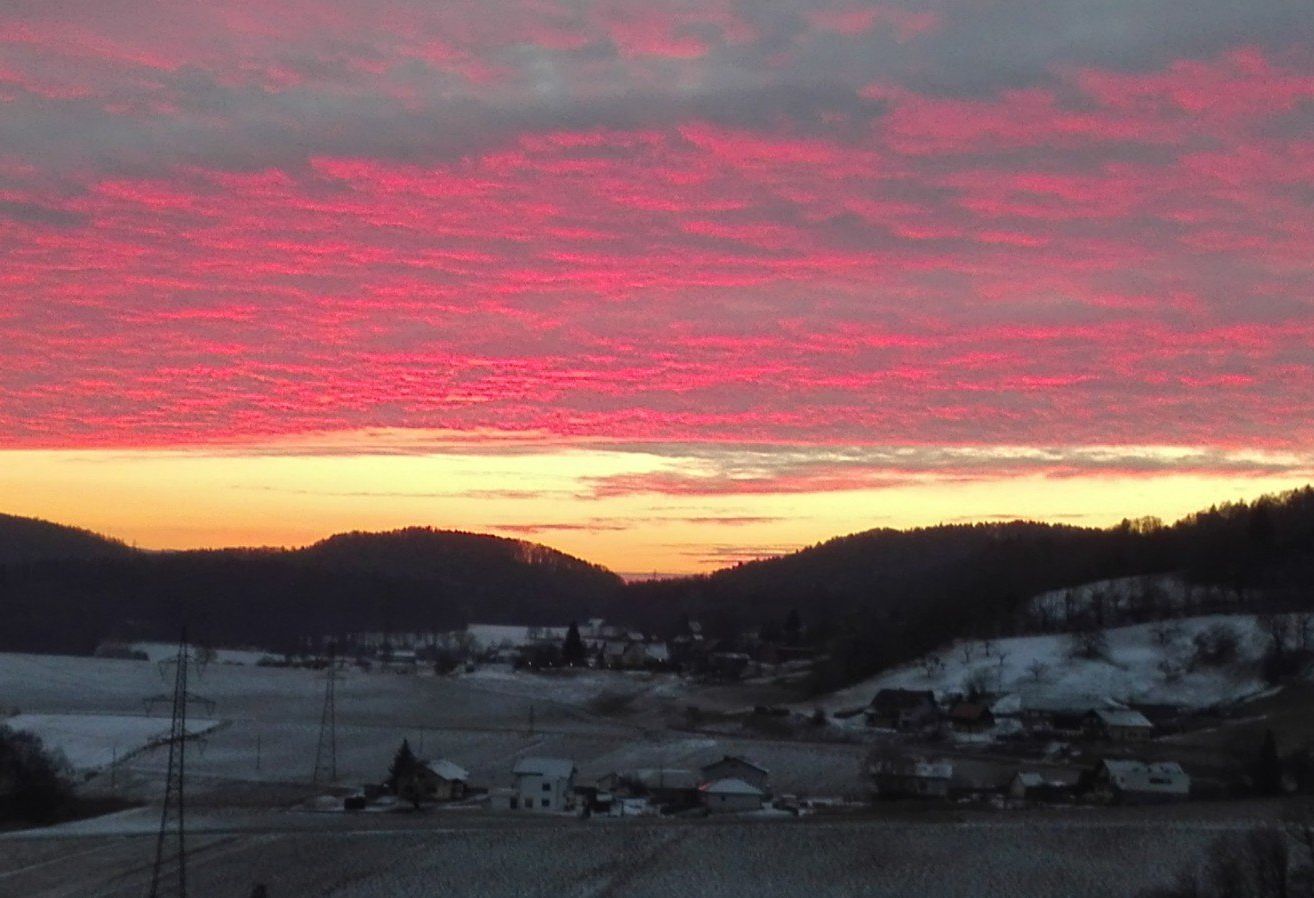 Ein Sonnenuntergangsblick auf eine verschneite Landschaft mit Bergen in der Ferne und einem kleinen Dorf darunter. Der Himmel ist in Rot-, Orange- und Gelbtönen gemalt.