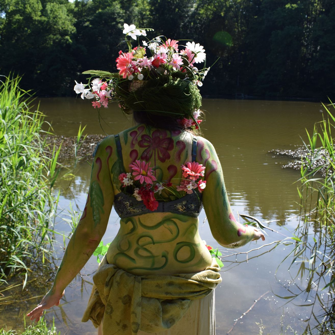 Eine Person mit einem Blumenkranz und Körperbemalung steht am Rand eines Teichs, geschmückt mit Blumen auf dem Rücken und den Schultern.