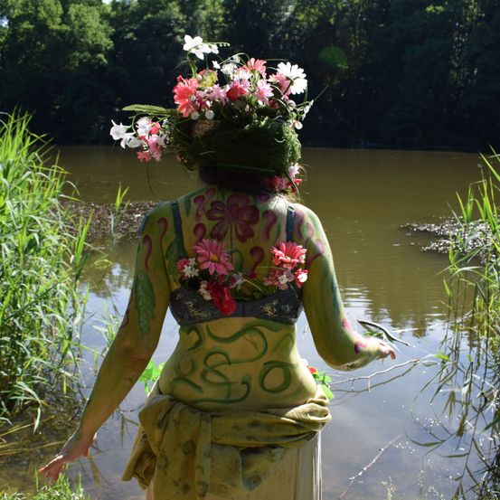 Eine Person mit einem Blumenkranz und Körperbemalung steht am Rand eines Teichs, geschmückt mit Blumen auf dem Rücken und den Schultern.