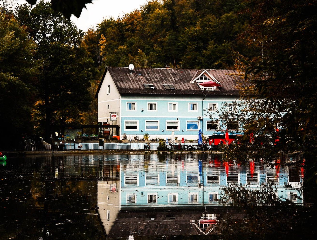 Ein blaues Haus mit schwarzem Dach steht am Fluss, umgeben von Bäumen und einem Zaun. Der Fluss spiegelt das Haus und die Bäume wider.