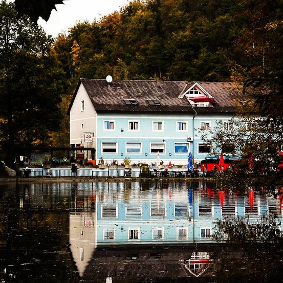 Ein blaues Haus mit schwarzem Dach steht am Fluss, umgeben von Bäumen und einem Zaun. Der Fluss spiegelt das Haus und die Bäume wider.