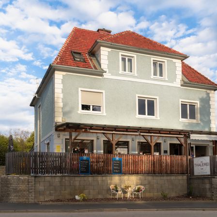 Ein zweistöckiges Haus mit rotem Dach und einer Holzzaun vorn. Es hat einen hölzernen Vorbau über dem Eingang und mehrere Fenster. Es gibt zwei blaue Schilder am Zaun und zwei Stühle mit einem kleinen Tisch davor.