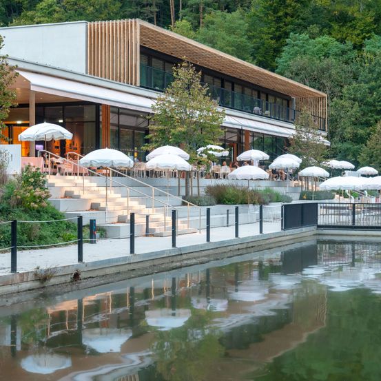 Ein modernes Gebäude mit Holzfassade und Glasfenstern befindet sich neben einem Kanal. Mehrere weiße Schirme sind auf den Stufen aufgestellt, die zum Eingang führen. Bäume und Pflanzen umgeben das Gebäude.