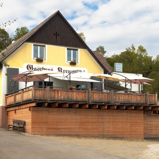 Ein gelbes und braunes Gebäude mit Balkon, mehreren Sonnenschirmen und einer Bank davor. Das Schild lautet 'Gasthaus Areuth'.