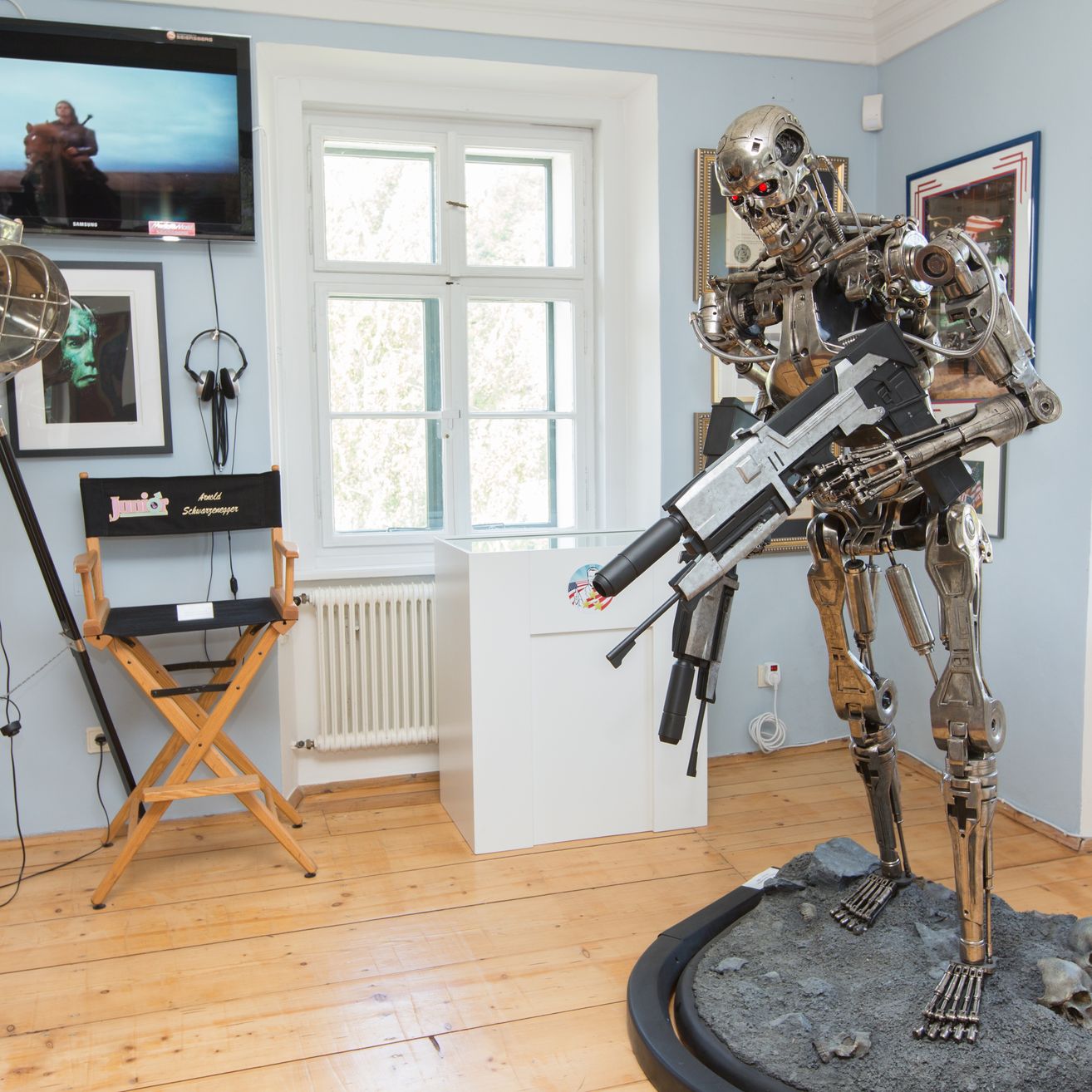 Ein Raum mit Holzboden, einer Terminator-Statue mit einer Waffe, einem Regisseurstuhl, einem Monitor und einem Heizkörper.