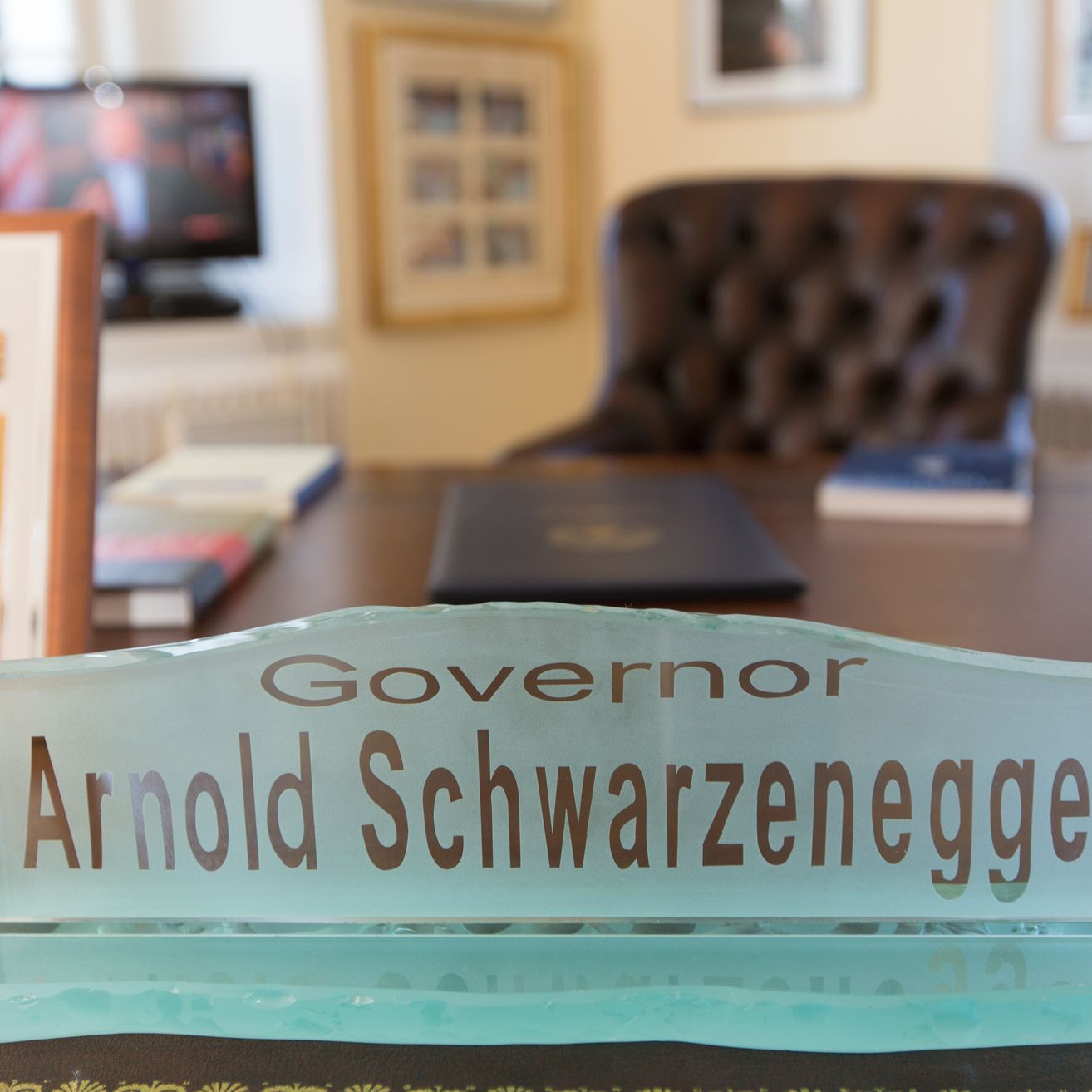 Ein Büroschreibtisch mit einem Glas-Namensschild, das 'Gouverneur Arnold Schwarzenegger' steht. Hinter dem Schreibtisch ist ein brauner Lederstuhl und ein Computermonitor. Gerahmte Bilder hängen an der Wand.