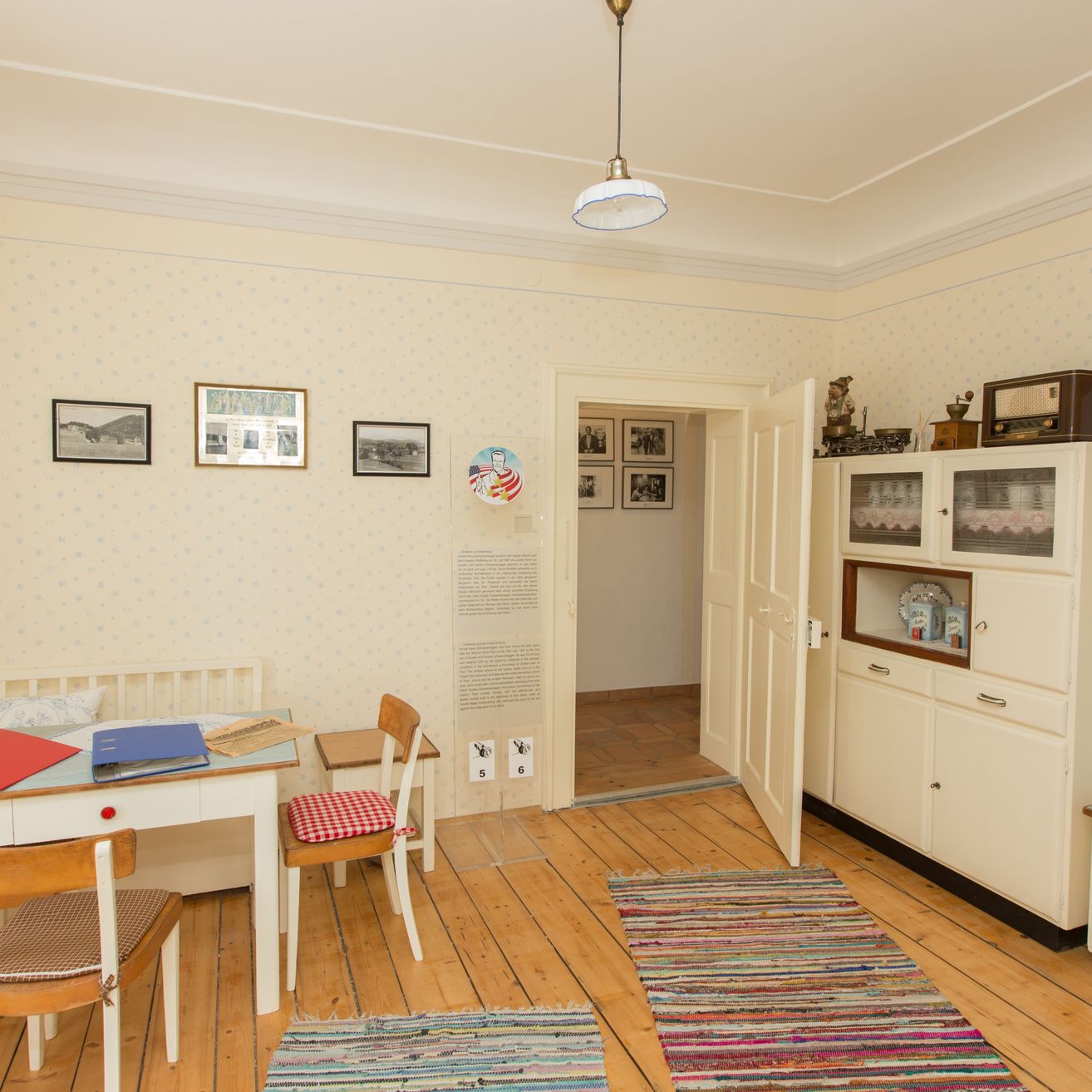 Ein Zimmer mit Holzböden und weißen Wänden. Ein Schreibtisch mit Büchern und einem roten Tuch steht in der Ecke. Ein Teppich liegt auf dem Boden. Ein Schrank mit Glastüren steht rechts. Eine Tür ist offen und führt in einen anderen Raum.