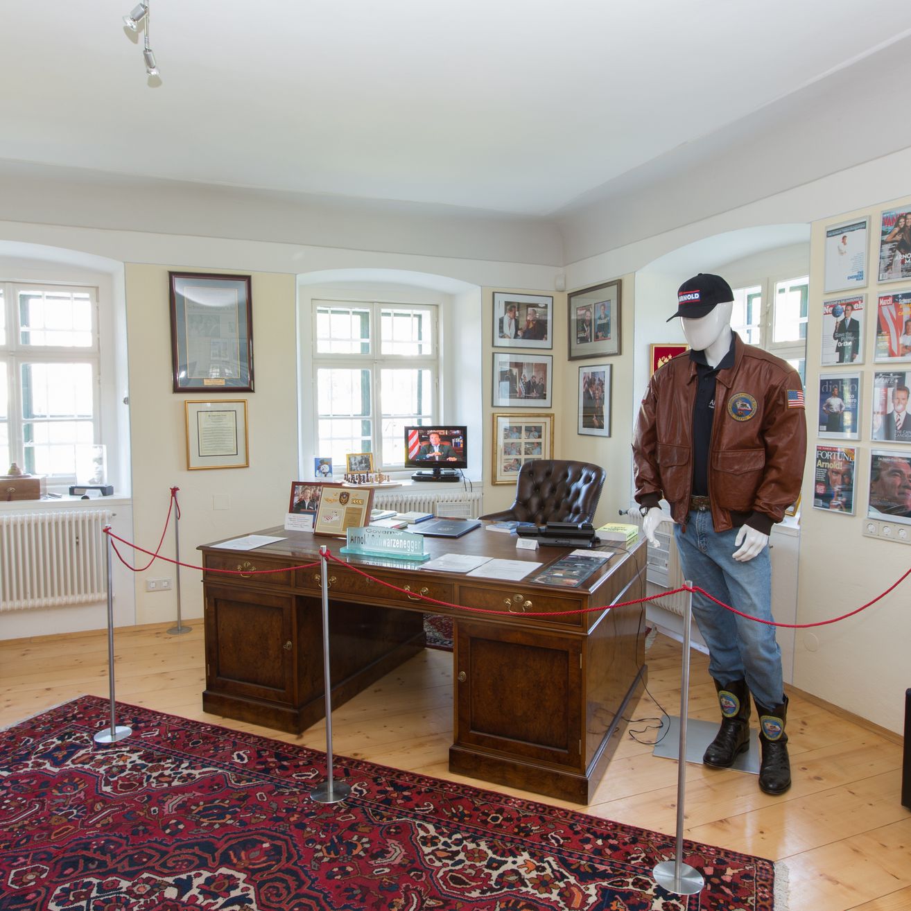Ein Raum mit einem Schreibtisch, einem Mannequin in einer Jacke und einem Teppich auf dem Boden. An der Wand befinden sich Bilderrahmen und eine Heizung.
